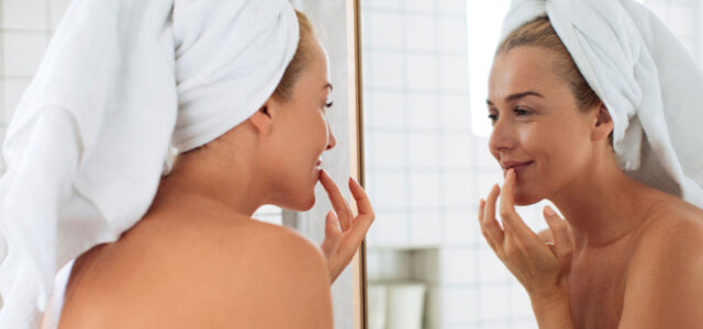 Frau mit Handtuch auf Kopf schaut sich in Spiegel an