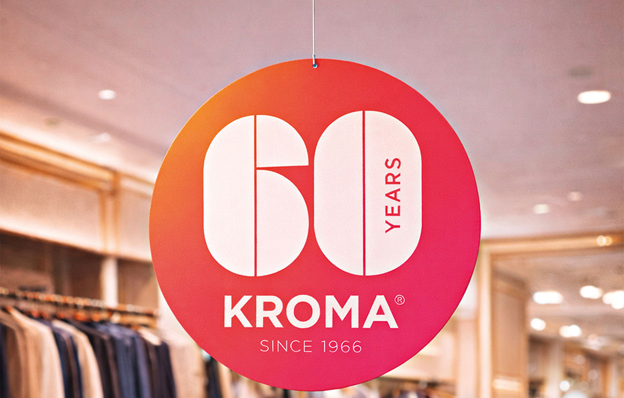 Runder Karton mit Aufschrift "60 Years Kroma" hängt an Decke. Gut ausgeleuchtes Kleidergeschäft im Hintergrund.