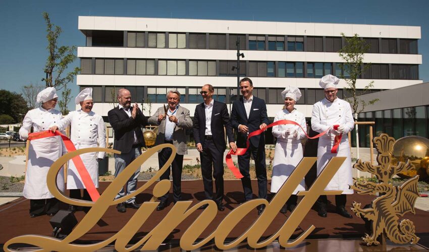 Personen in Anzügen und Kochkleidung zerschneiden gemeinsam Band vor Schritzug "Lindt"