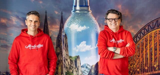 Thomas Horak und Wolfgang Graski vor Bild mit großer Almdudler-Flasche und Skyline von Köln