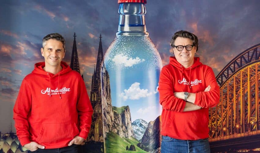 Thomas Horak und Wolfgang Graski vor Bild mit großer Almdudler-Flasche und Skyline von Köln